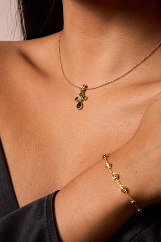 Bendita cross pendant necklace in gold with emerald-green stones and central pink gem, paired with matching jewelry.

Dije Bendita en forma de cruz con baño de oro, piedras verde esmeralda y piedra rosa central.