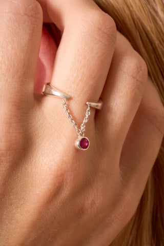Close-up of silver ring with red gemstone chain detail on hand.

Primer plano de anillo de plata con detalle de cadena y gema roja en la mano.