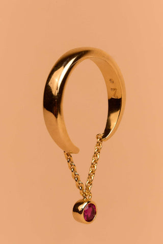 Cosma gold ring standing upright, showing its open band linked with a chain and red bezel-set stone.

Anillo Cosma en oro de pie, mostrando su aro abierto unido con una cadena y piedra roja.